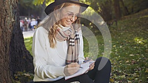 Close up shot of young woman sitting on the grass in the city park and writing some information in note book.