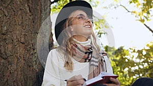 Close up shot of young woman in stylish hat writing something in notebook while relaxes in the city park.