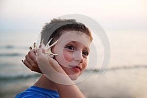 Close up side portrait of a beautiful young boy on holiday holding a sea shell to his ear and smiling, listening to the sound of