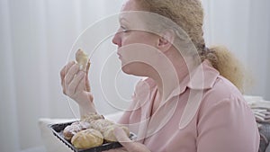 Close-up side view of chubby senior woman chewing sweet bun. Portrait of redhead Caucasian lady having obesity eating