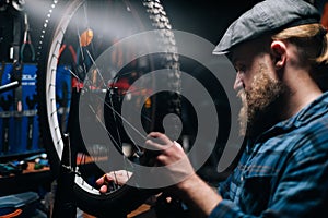 Close-up side view of concentrated bearded repairman checking bicycle wheel spoke with bike spoke wrench in bike repair