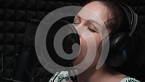 Close up side view of face of attractive brunette girl is recording voice to microphone in music studio with black background. Clo