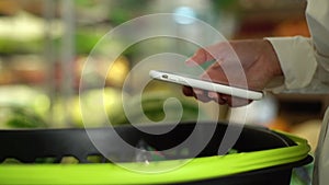 Close-up side view hands of unrecognizable young woman customer browsing on smartphone under grocery cart in supermarket