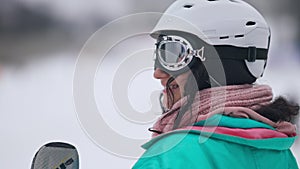 Close-up side view of happy smiling young woman in ski goggles and helmet looking away standing at winter resort with