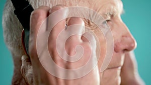 Close-up side view of senior male putting headphones on head over blue background. Elderly man puts earphones on head.