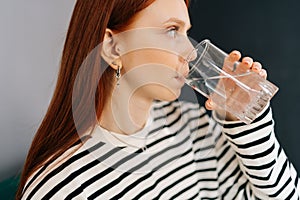 Close-up side view of thirsty redhead female holding glass, drinking still water enjoy natural aqua at home, preventing