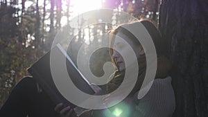 Close up side view of young charming woman in cozy clothes holding book in hands and reading in sun rays at fall forest