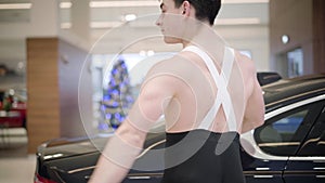 Close-up side view of young male caucasian ballet dancer bending back and spinning moving along car in auto showroom