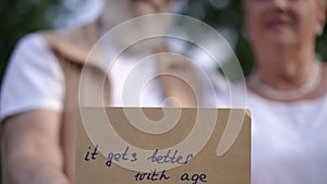 Close-up of a sign saying it gets better with age, held in hand by an elderly couple while standing on a path in a city