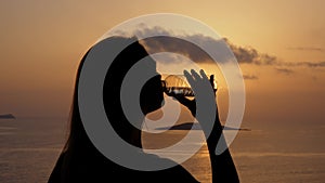 Close up silhouette of unrecognizable girl drink water from transparent bottle at sunset, in background ocean or sea