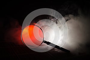 Close up Single Magnifying Glass with Black Handle, Leaning on the Wooden Table on orange red smoke dark background. Surreal backg