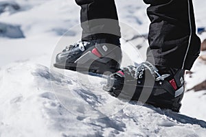 A close-up of a skier`s leg in ski boots without skis stands on a knoll of bliss against the background of mountains on