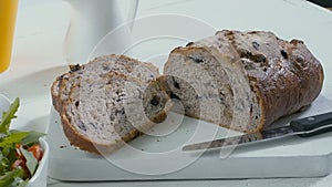 Close-up of sliced rye raisin bread on the white cutting board. There are also fresh veggies, white milk jug and yellow mug on the