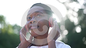 Close-up slim charming African American woman in headphones smiling listening to music standing in sunbeam. Headshot of
