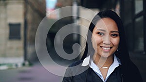 Close-up slow motion portrait of attractive Asian girl looking at camera with happy smile standing in the street wearing