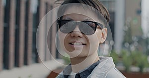 Close-up slow motion portrait of cheerful Asian man wearing stylish sunglasses smiling looking at camera outdoors in