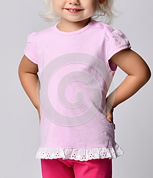 Close-up of small blonde baby girl in pink stylish t-shirt with ruffles standing holding hands at waist