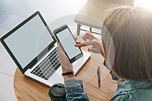 Close-up of smartphone with blank screen in hands of young woman sitting at round wooden table and touching screen