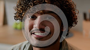 Close up smiling Arabian man homeowner with curly hair vlogger look at camera at kitchen. Portrait Indian male face with
