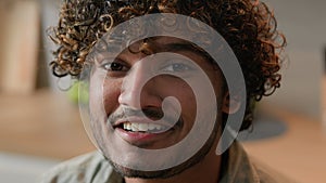 Close up smiling Arabian man homeowner with curly hair vlogger look at camera at kitchen. Portrait Indian male face with