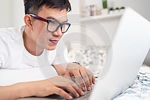 Close up of smiling Asian man wearing glasses using laptop computer working from home laying on bed, digital lifestyle social