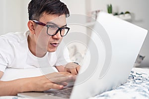 Close up of smiling Asian man wearing glasses using laptop computer working from home laying on bed, digital lifestyle social