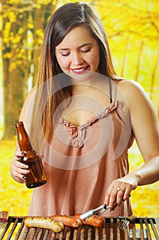 Close up of smiling beautiful young woman holding a bottle in one hand and using a tongs and turning the grilling