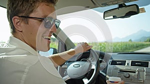 CLOSE UP: Smiling Caucasian man holds a takeaway cup of coffee while driving.