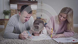 Close-up of smiling Caucasian man and woman kissing little girl on cheeks simultaneously. Cute brunette daughter drawing