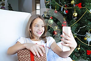 Close up smiling caucasian young preteen girl talking on phone, sitting near festive Christmas tree at home, happy