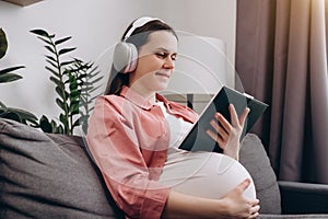 Close up of smiling cute young pregnant woman listen to music with wireless headphone and reading book to tell story to unborn