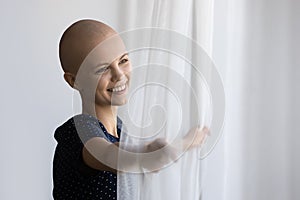 Close up smiling dreamy hairless woman opening curtains in morning