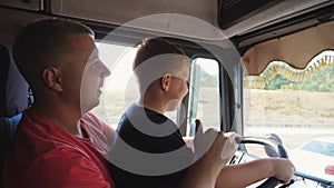 Close up of smiling ginger child holds the big steering wheel sitting on lap of father and learning to drive the truck