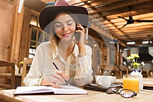 Close up of a smiling girl in hat sitting at the cafe table indoors, talking on mobile phone