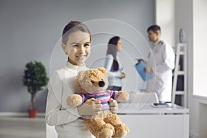 Close up of smiling healthy little girl hugging soft toy bear in doctor& x27;s office looking at camera.