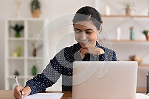 Close up smiling Indian woman taking notes, using laptop