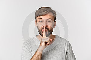 Close-up of smiling man in gray t-shirt, hushing, telling a secret, standing over white background