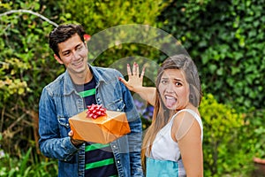 Close up of smiling man holding a gift and his girlfriend, stretching her arm ignoring him and woman doing a disgusting