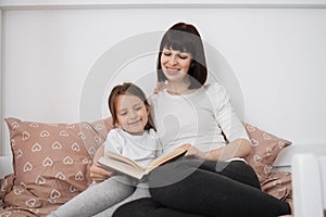 Close up of smiling mom and little daughter relax in cozy bed read funny interesting children book