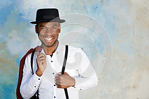 Close up smiling young african american male fashion model posing with suspenders and hat by wall