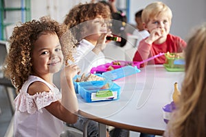 Close up of smiling young children sitting at a table eating their packed lunches together at infant school, girl smiling to camer