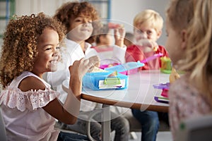 Close up of smiling young children sitting at a table eating their packed lunches together at infant school, selective focus