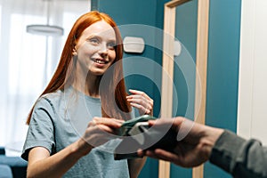 Close-up of smiling young woman customer making contactless payment using smartphone using POS wireless terminal to