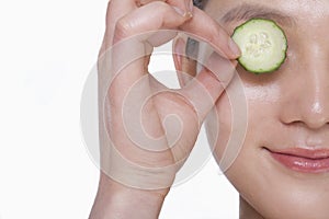 Close up of smiling young woman holding up a cucumber slice over her eye, studio shot