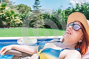 close-up of a smiling young woman wearing a hat and sunglasses lying in a hammock sunbathing by a hotel pool. girl