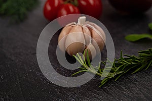 Close-up of a sprig of rosemary on a metallic dark background, with a head of garlic and other vegetables in the background