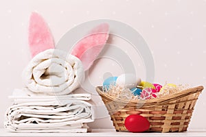 A close-up of a stack of clean white bedding, towels and Easter bunny ears, a basket of eggs on a table.
