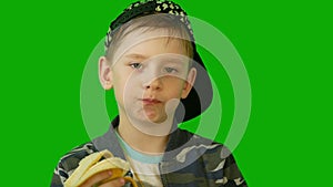 Close-up and a starving European boy in a cap eats a banana against a green background.