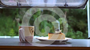 Close-up steaming hot coffee or tea cup with cake on table with view on green forest. Coffee cup with smoke in camper