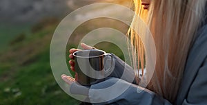 Close-up of steel mug with hot drink in hands of young blonde girl at susnet.